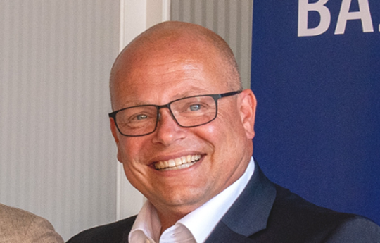 A man with a bald head and black glasses looks into the camera, smiling.