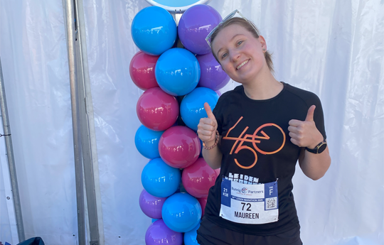 A woman in a blue T-shirt printed “450” raises both thumbs up. In the background blue, purple and pink balloons.
