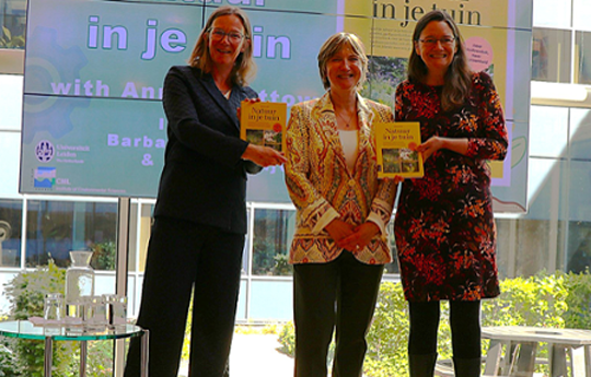 Three women on a stage. The two women on either side both hold up a book.